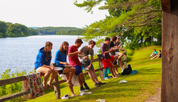 Students eat, leaning on a wood fence