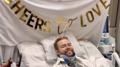 Franz Wall in hospital bed and neck brace, with "Cheers to Love" sign above him