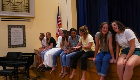 A group of women sits on the edge of stage, along with Fr. Sebastian Walshe.