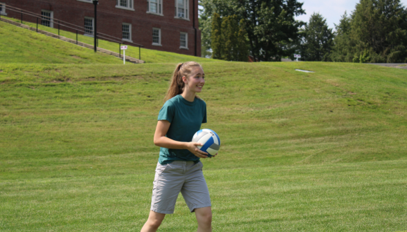 A student holding the volleyball, afront the green hillside to Gould