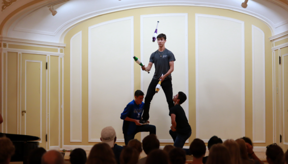 A student juggles pins while being standing, held up by two other students