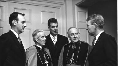 Black and white photo of 5 men in suit and robes talking